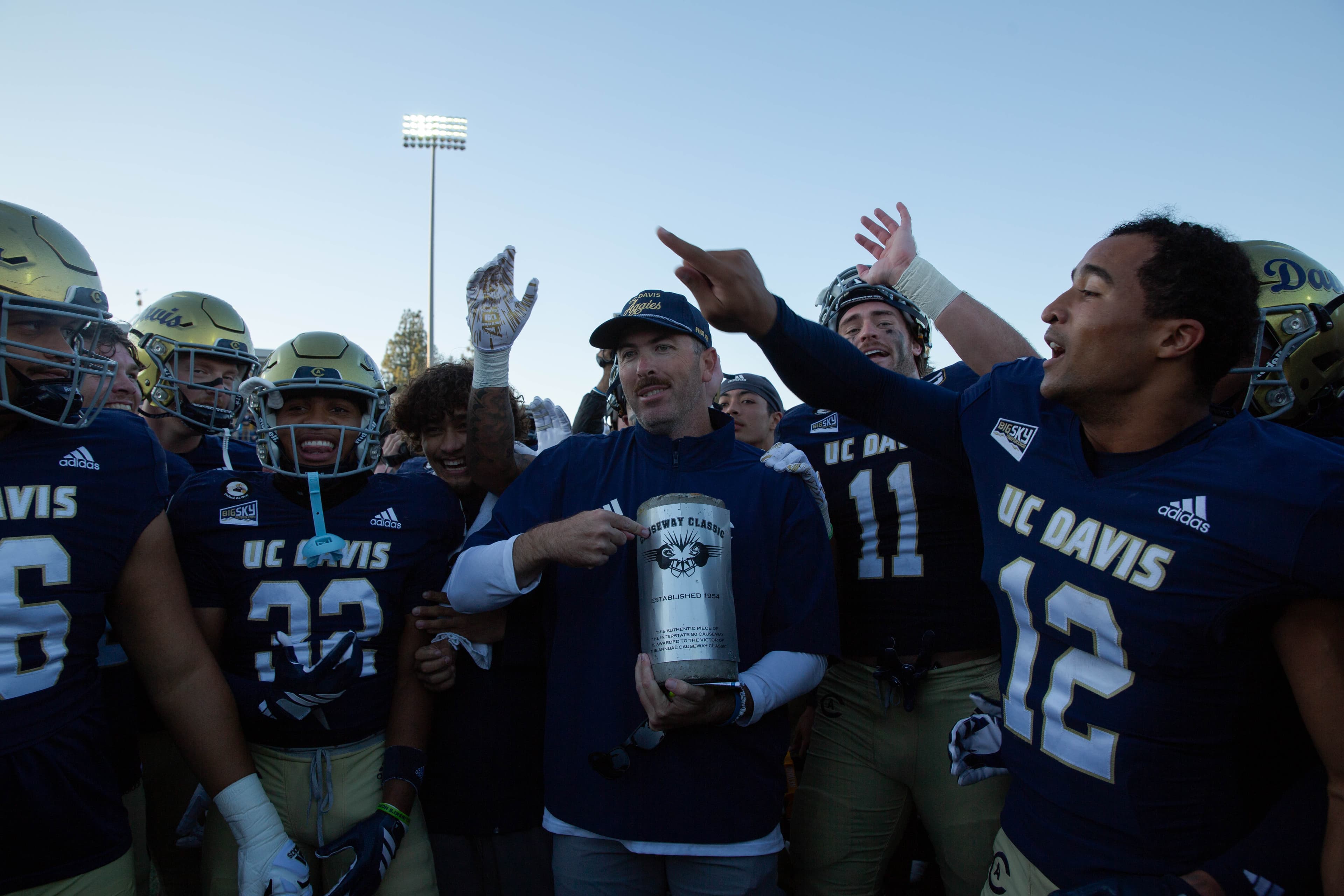 UC Davis football players gather around their head coach, who holds a silver trophy.