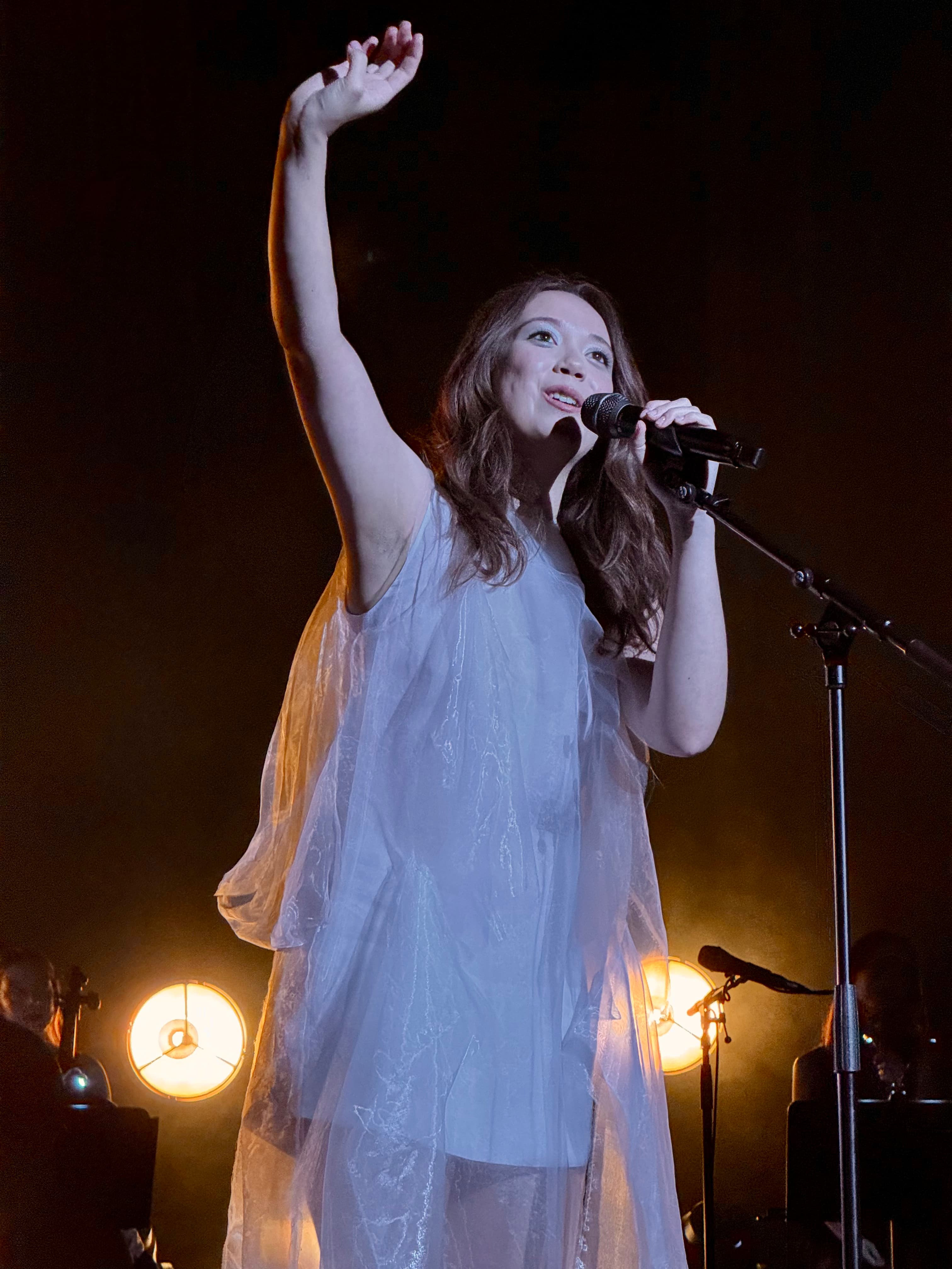 Singer performing on stage with a microphone and raised hand, warm lights glowing in the background ( LAUFEY / AGGIE?)
