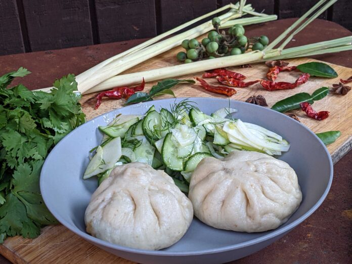 Bao with a cucumber salad on a concave plate on a table with cilantro and dried pepper